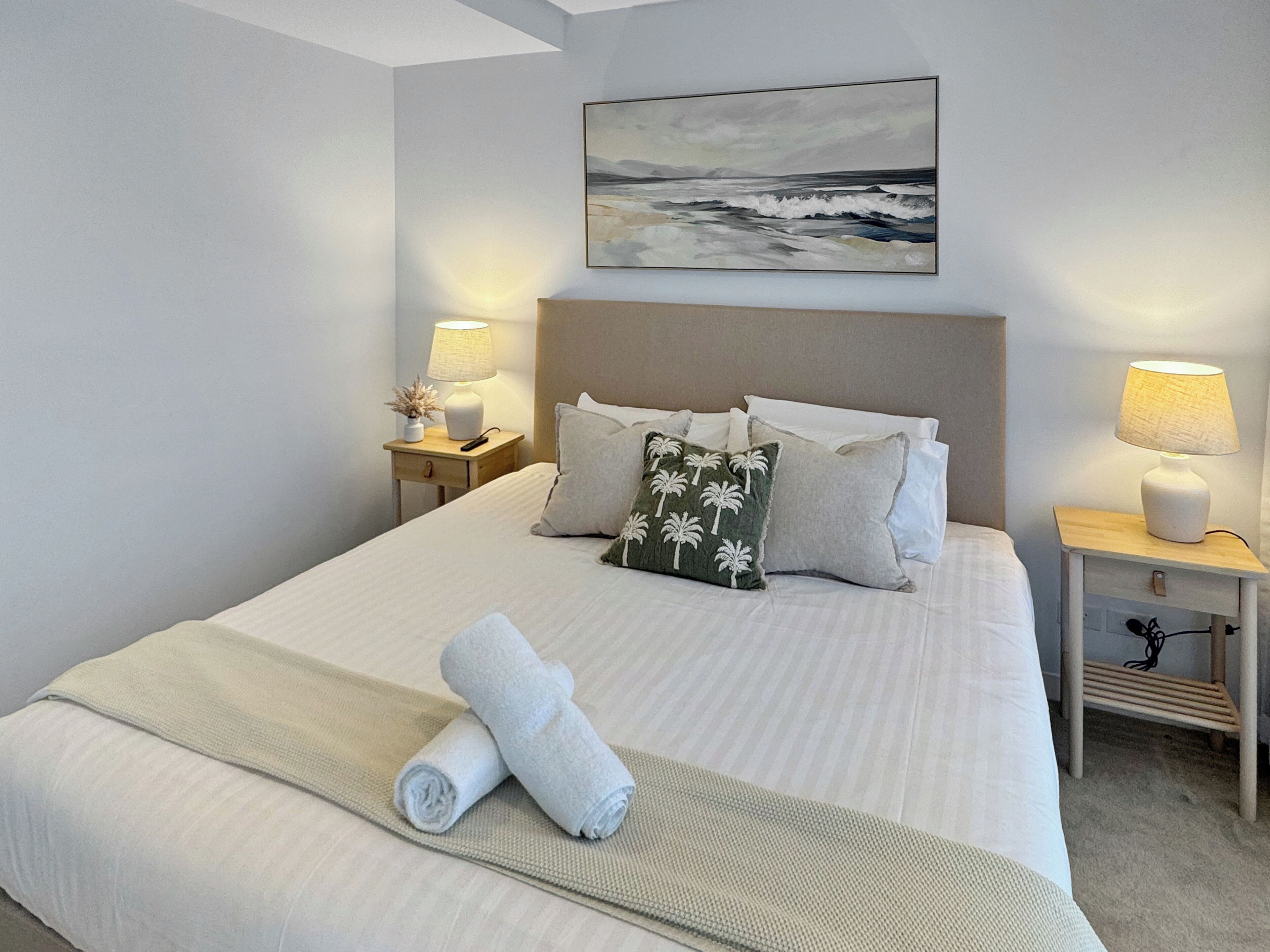 Un lit bien fait, avec du linge de lit blanc, une serviette enroulée et un oreiller orné de motifs de palmiers. Il y a deux tables de chevet avec des lampes et, au-dessus du lit, un tableau représentant un paysage.