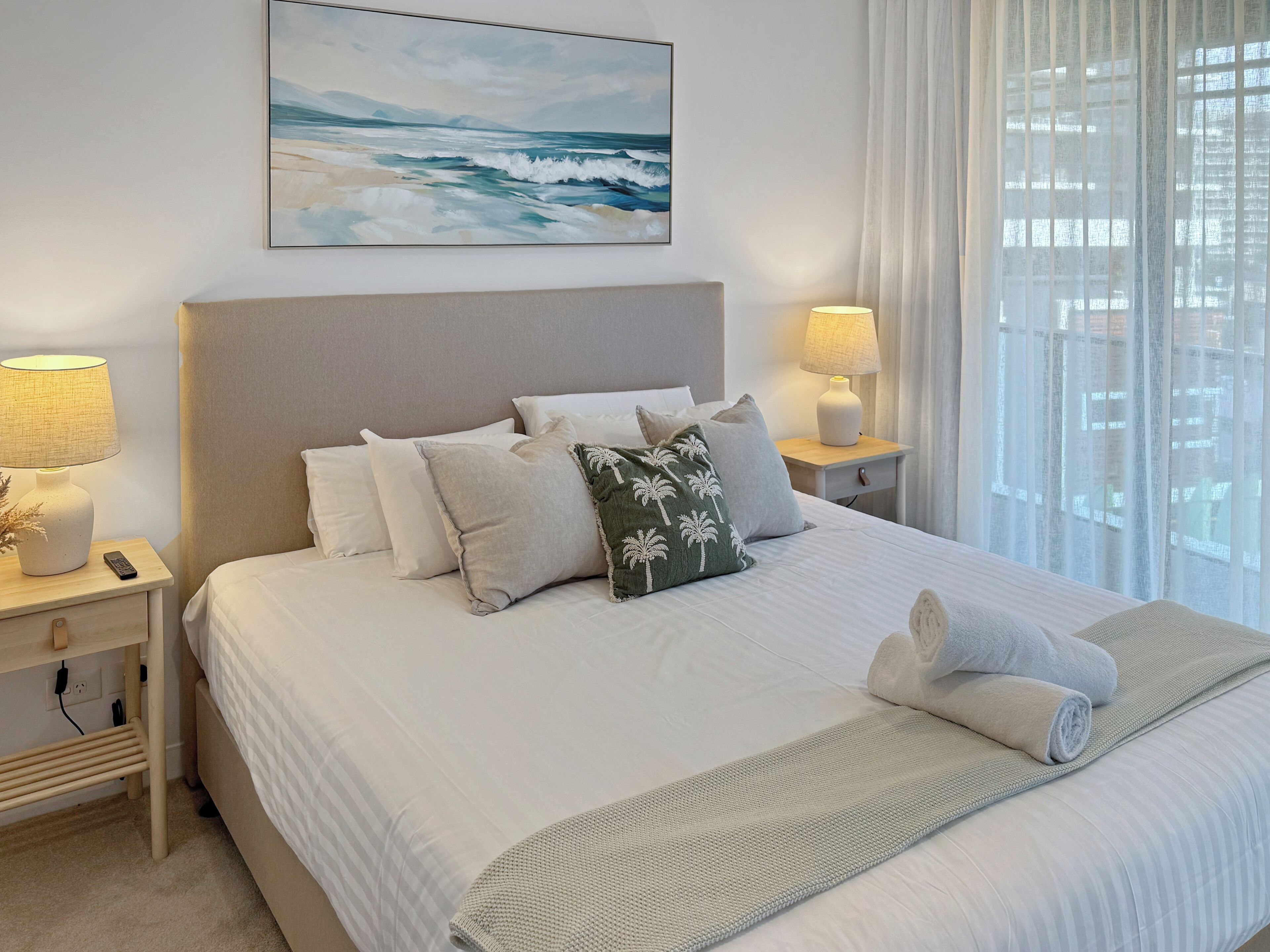 A neatly made bed with white bedding and a palm tree pillow, a bedside lamp, and a painting of a beach scene.