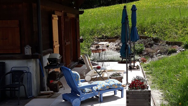 Ein überdachter Sitzbereich mit Kieshof, einem Holzgebäude und einem Garten mit Blumen und Bäumen im Hintergrund.