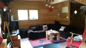 Ein gemütliches Blockhaus-Interieur mit Holzboden, einem dunkelbraunen Sofa, einem kleinen Holztisch, einem roten Sessel und einem großen Fenster, das für natürliches Licht sorgt.