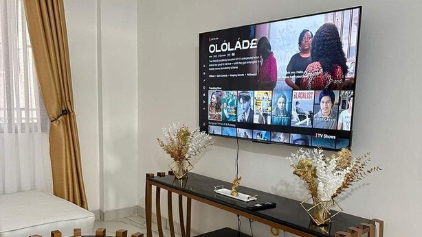 A modern living room with a TV mounted on the wall displaying a show, a round coffee table surrounded by chairs, and a sofa. - Lux Suites Narayan Apartments Kisumu (Kisumu)