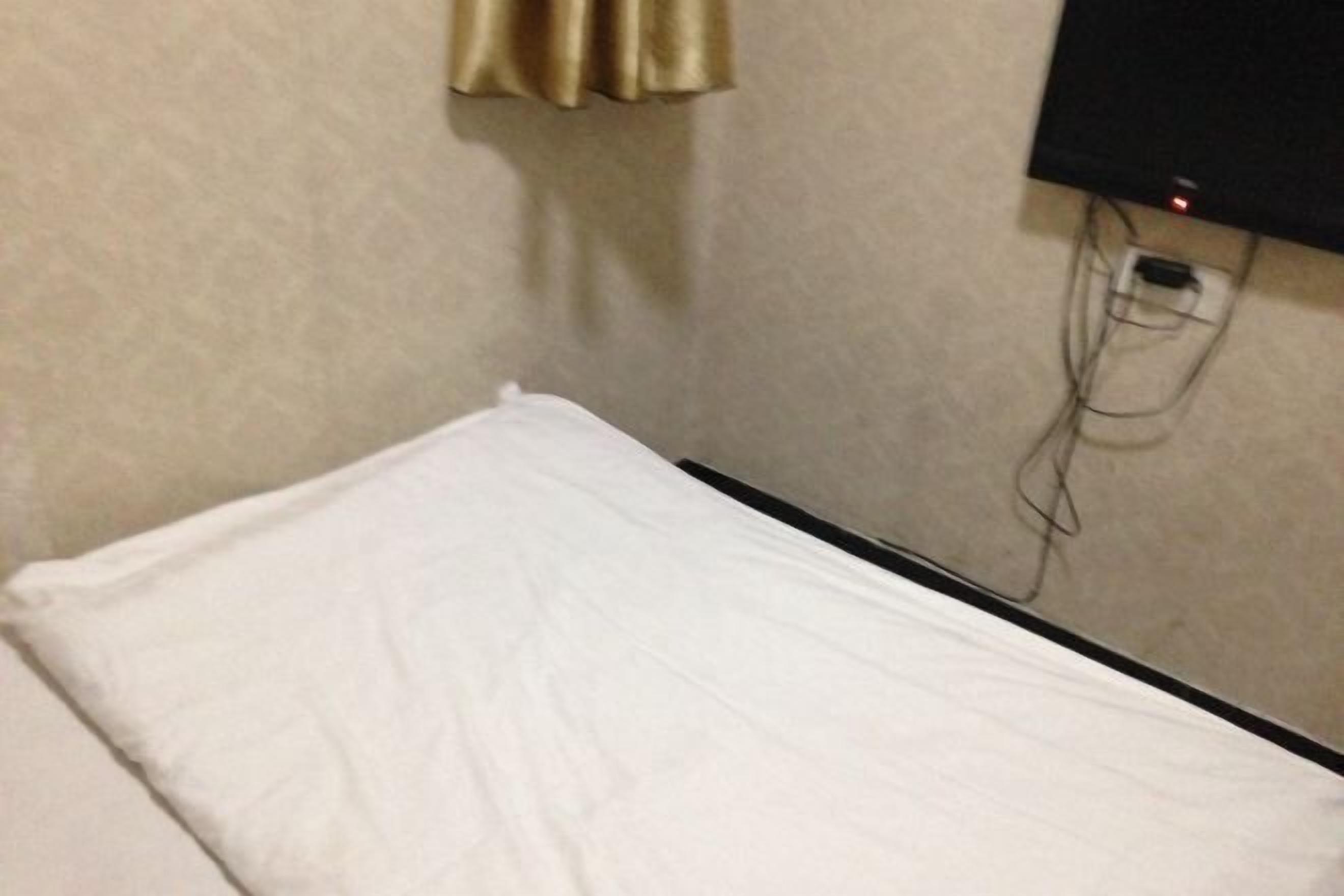 A small, single-bed room with a white bedspread, a television mounted on the wall, and visible electrical outlets.