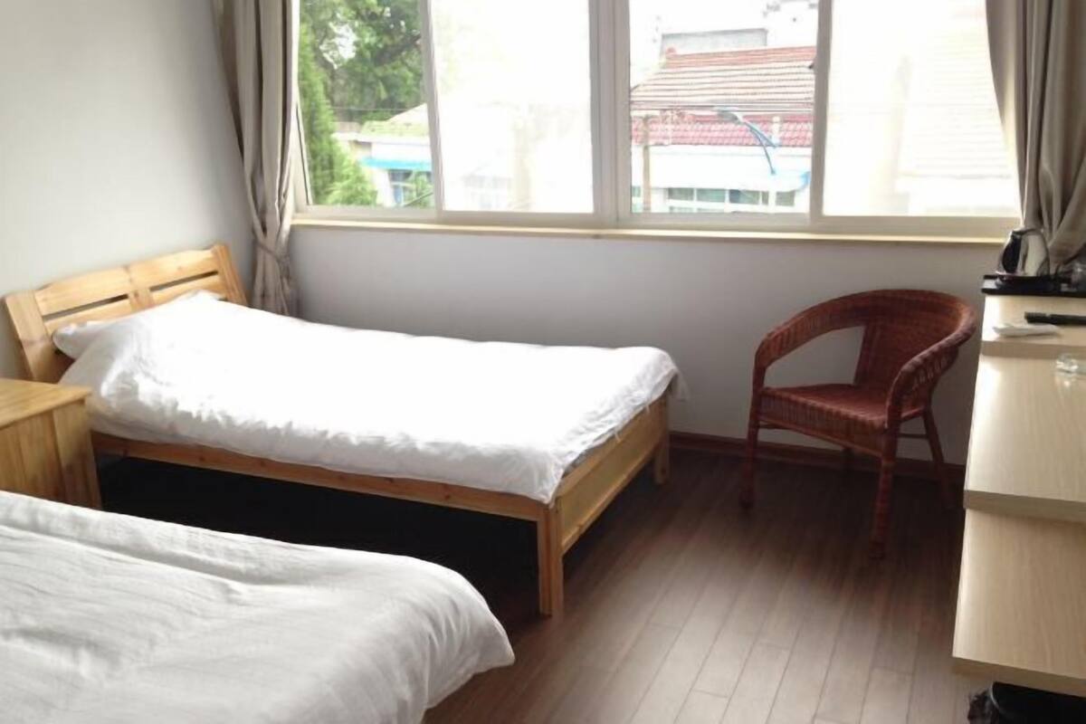 A bedroom with two single beds, a wooden headboard, a red wicker chair, a desk with a lamp, and a window with curtains.