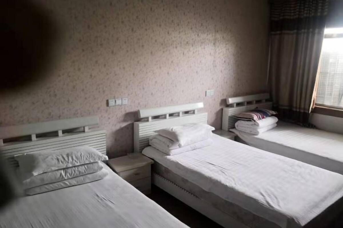 A hotel room with three single beds, a nightstand, a wall-mounted outlet, and a window with curtains.