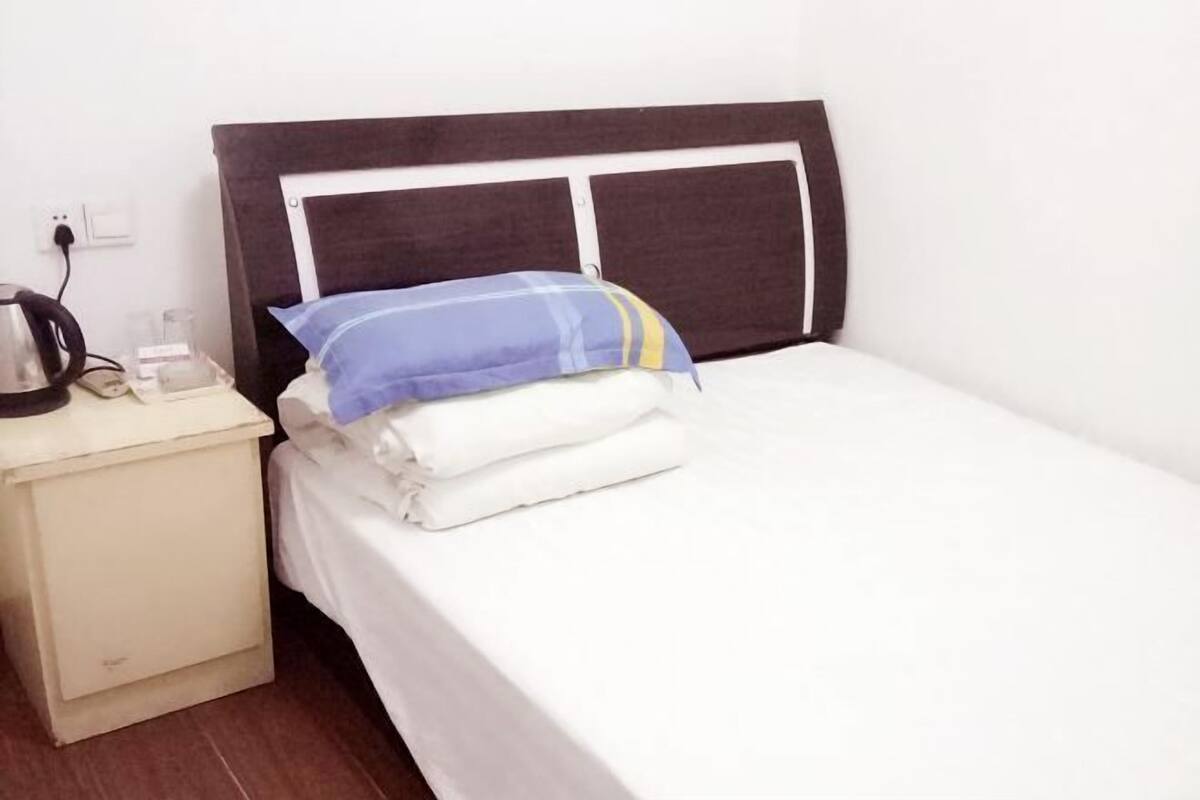 A bed with a white mattress and a blue and white striped pillow, a bedside table with a kettle, and a power outlet.
