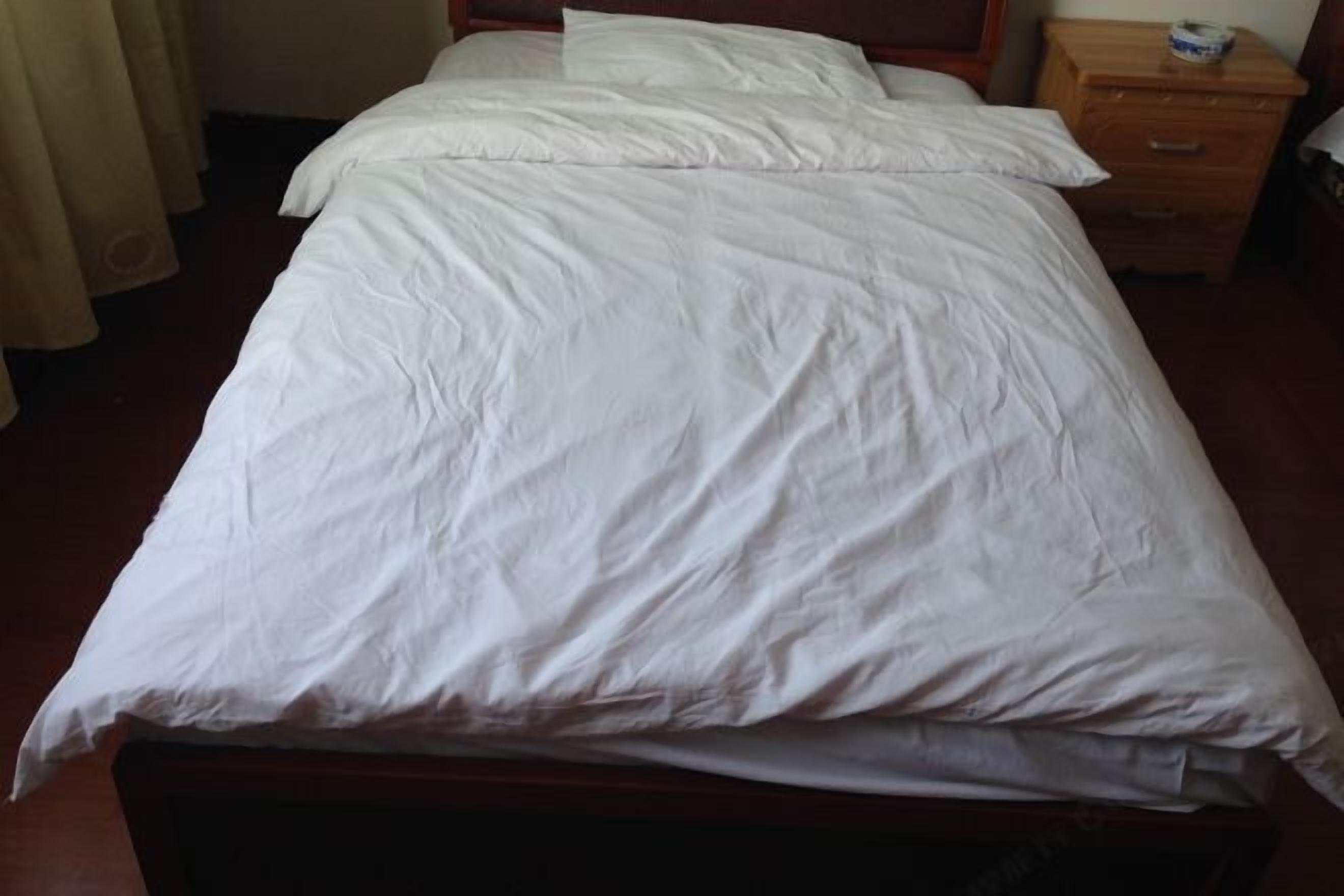 A neatly made bed with white bedding, a wooden headboard, and a wooden nightstand with a small item on it.