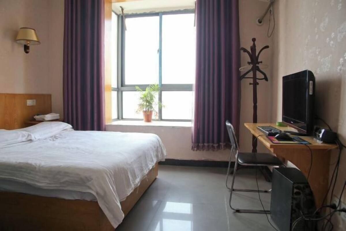 A hotel room with a bed, a desk with a computer, a chair, a TV, and a window with curtains.
