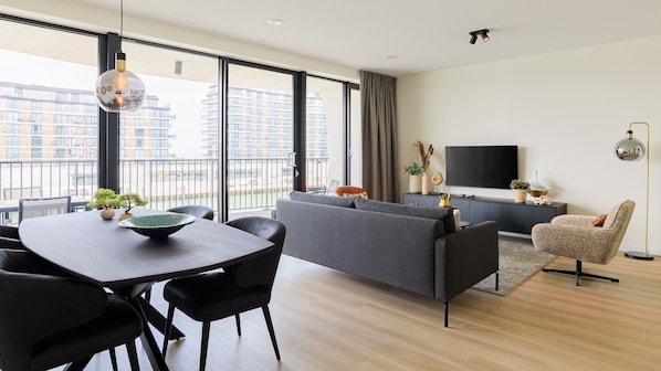 Una sala de estar moderna con sofá, mesa de comedor, sillas, televisor y balcón con vista a edificios.