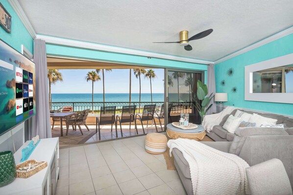 A room with a TV displaying streaming services, a balcony with a view of the ocean, a ceiling fan, and a sofa with cushions.