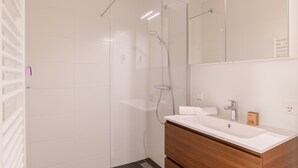 Moderni kylpyhuone, jossa on lasinen suihkuseinä, valkoinen pesuallas ja puinen allaskaappi.
