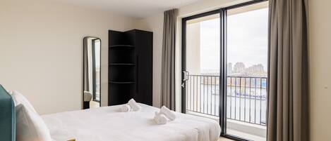 A modern hotel room with a large bed, a black wardrobe, a mirror, and a view of the city through a sliding glass door.