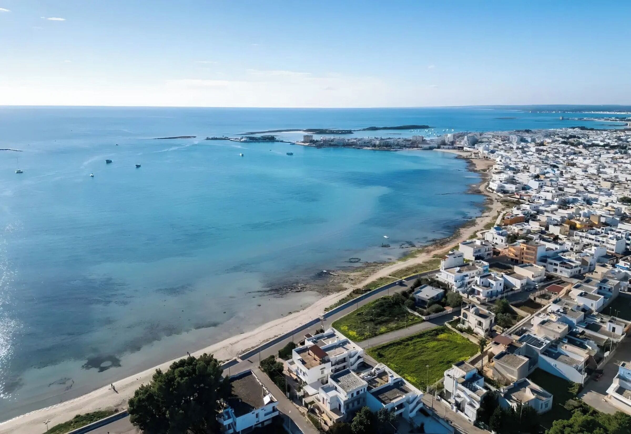 An aerial view of a coastal town with a beach, clear blue waters, and densely packed white buildings.