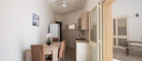 Cocina compacta con microondas, estufa y gabinetes. Hay una mesa de comedor con sillas y un ventilador de techo. Una puerta corrediza de vidrio da a un área exterior con una banca.