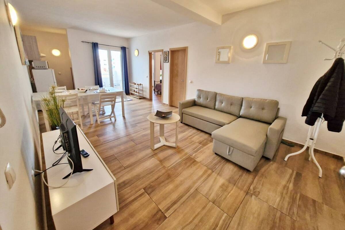 Ein modernes Wohnzimmer mit einer Couch, einem kleinen Tisch und einem Fernseher auf einem Schrank. Im Hintergrund ist ein Essbereich mit Tisch und Stühlen zu sehen.