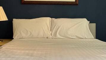 Una cama con sábanas blancas, un cuadro enmarco de un paisaje con caballos y una lámpara sobre la mesita de noche.