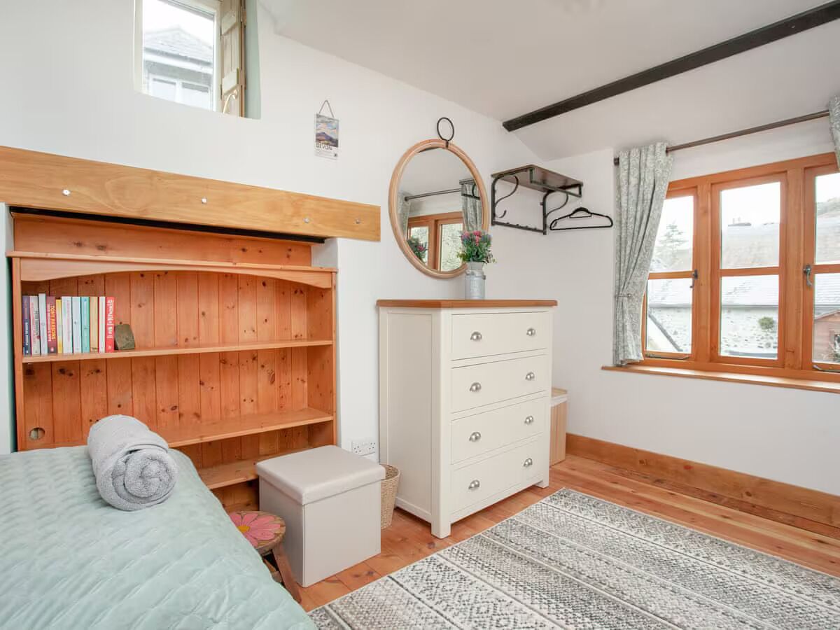 A bedroom with a bed, a wooden bookshelf, a dresser, a mirror, and a window with curtains.
