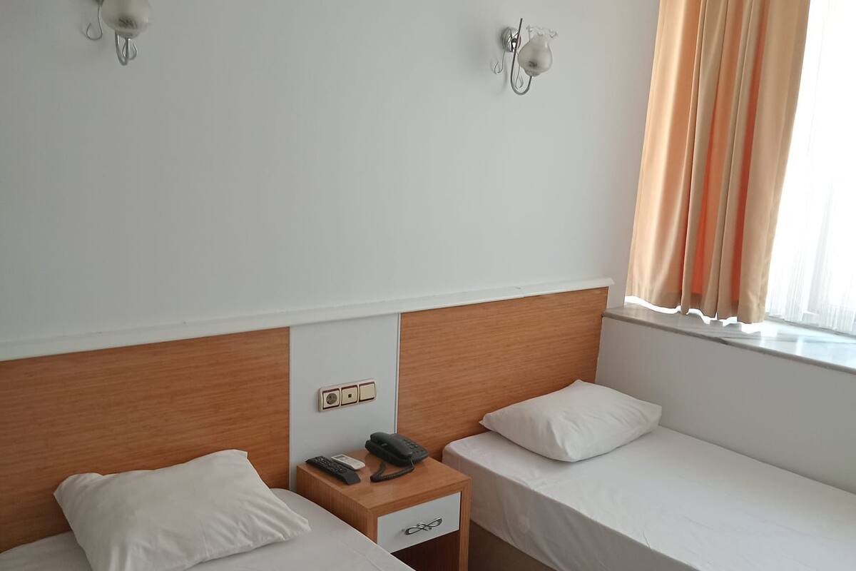 A hotel room with two single beds, a wooden headboard, a bedside table with a phone and a lamp, and a window with curtains.