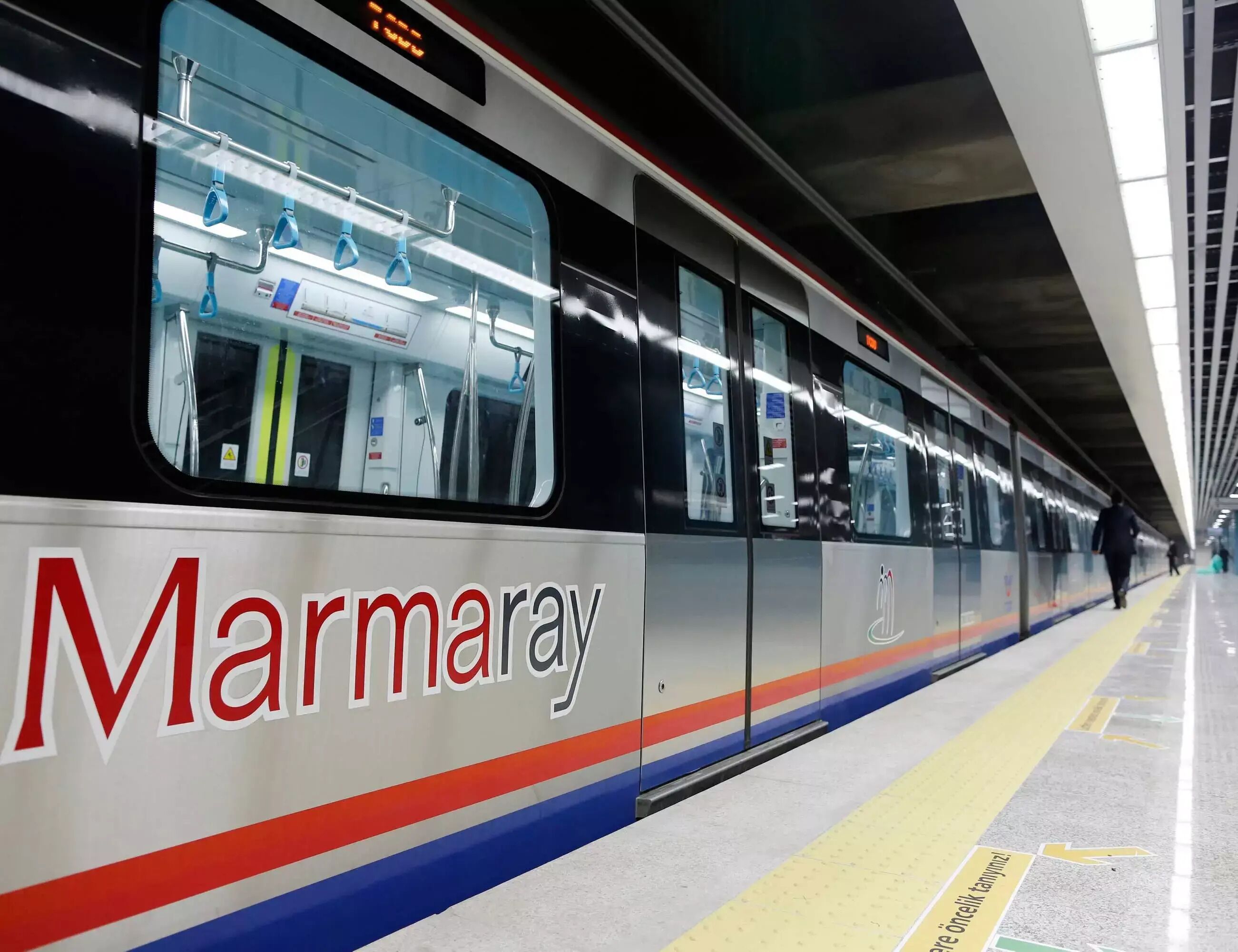 A train with 'Marmaray' branding, a platform with yellow safety lines, and a person walking in the distance.