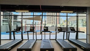 Een sportschool met vijf loopbanden op een rij, grote ramen en uitzicht op gebouwen.