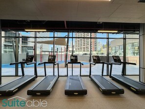 Un gimnasio con cinco cintas de correr alineadas, ventanas amplias y vistas a edificios.