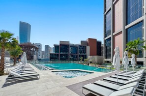 Uma área de piscina no terraço com espreguiçadeiras e guarda-sóis, cercada por edifícios modernos e palmeiras.