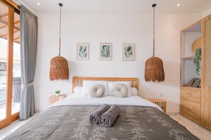 Een net opgemaakt bed met een grijze bloemenovertrek en opgerolde handdoeken, een houten hoofdbord en een groot raam met gordijnen.