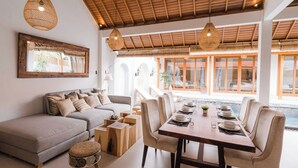 A modern living room with a sofa, dining table, and chairs, a large window, and a ceiling with wooden beams. - Villa Niji - 3BR Villa with Pool Seseh (Munggu)