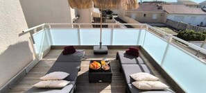 Eine Dachterrasse mit zwei Liegestühlen, einem Tisch mit Früchten und einer Flasche sowie einem Strohdachschirm.