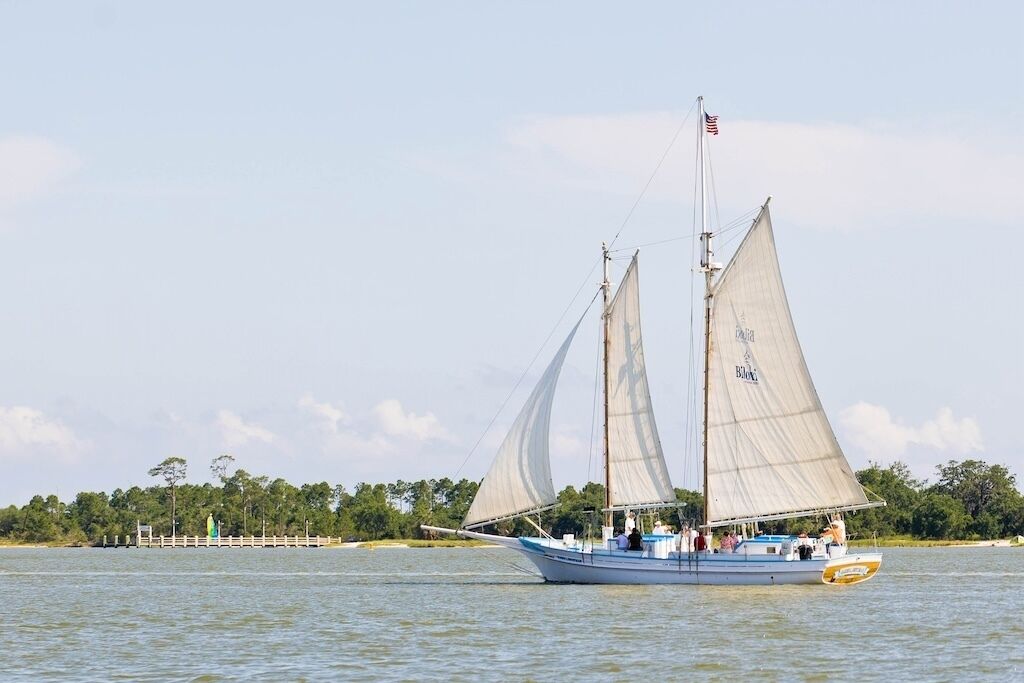A sailboat with multiple sails on a body of water, surrounded by a dock and trees.