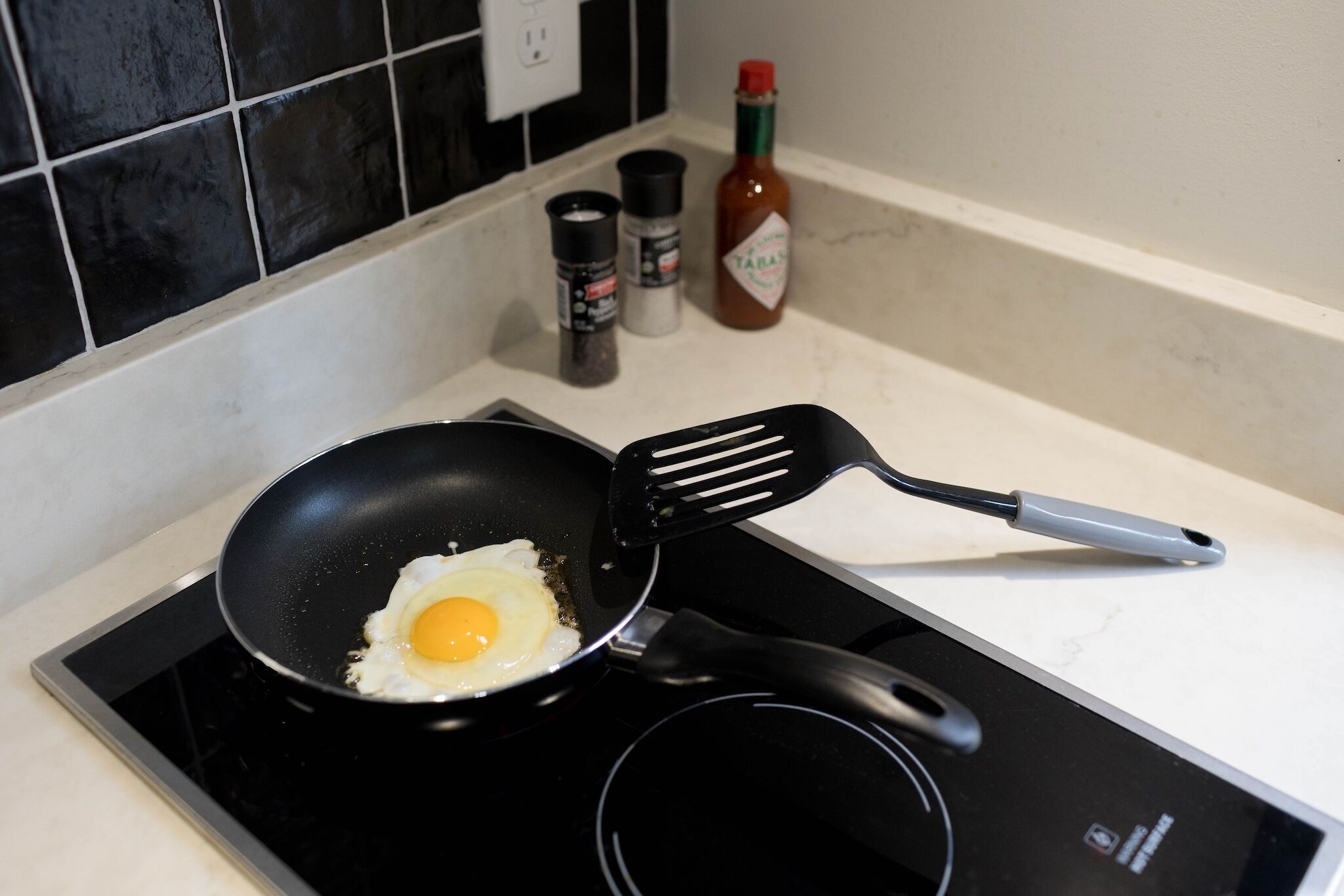 Een keuken met een koekenpan op een inductiekookplaat, een spatel en kruiden op het aanrecht.