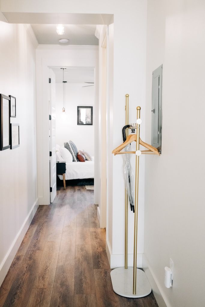 A hallway with a wooden floor, a coat rack with a white base, and a bed in the background.