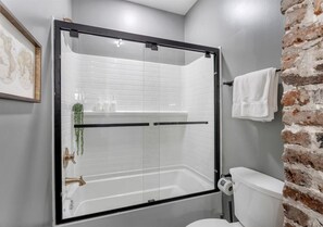 Un baño moderno con ducha envuelta en vidrio, paredes de azulejos blancos, una pared de ladrillo decorativa y un inodoro con un rollo de papel higiénico.