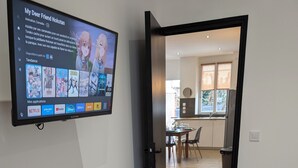Une chambre moderne équipée d’un téléviseur diffusant une émission, d’une télécommande, d’une commode blanche et d’une porte en verre donnant sur un coin repas.