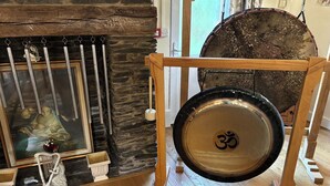 A room with a stone fireplace, a hanging metal wind chime, a drum with an Om symbol, and a statue of a deity. - Peaceful Cottage in Sound Healing Retreat (Cardigan)