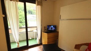 A room with a wooden floor, a television on a wooden cabinet, a wicker chair with a red cushion, and a sliding glass door leading to an outdoor area. - Luchon - Studio 2/3 pers., balcony, cable car, near shops and thermal baths (Bagnères-de-Luchon)