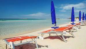 Een strand met rijen ligbedden en blauwe parasols, helder turquoise water en een zandstrand.