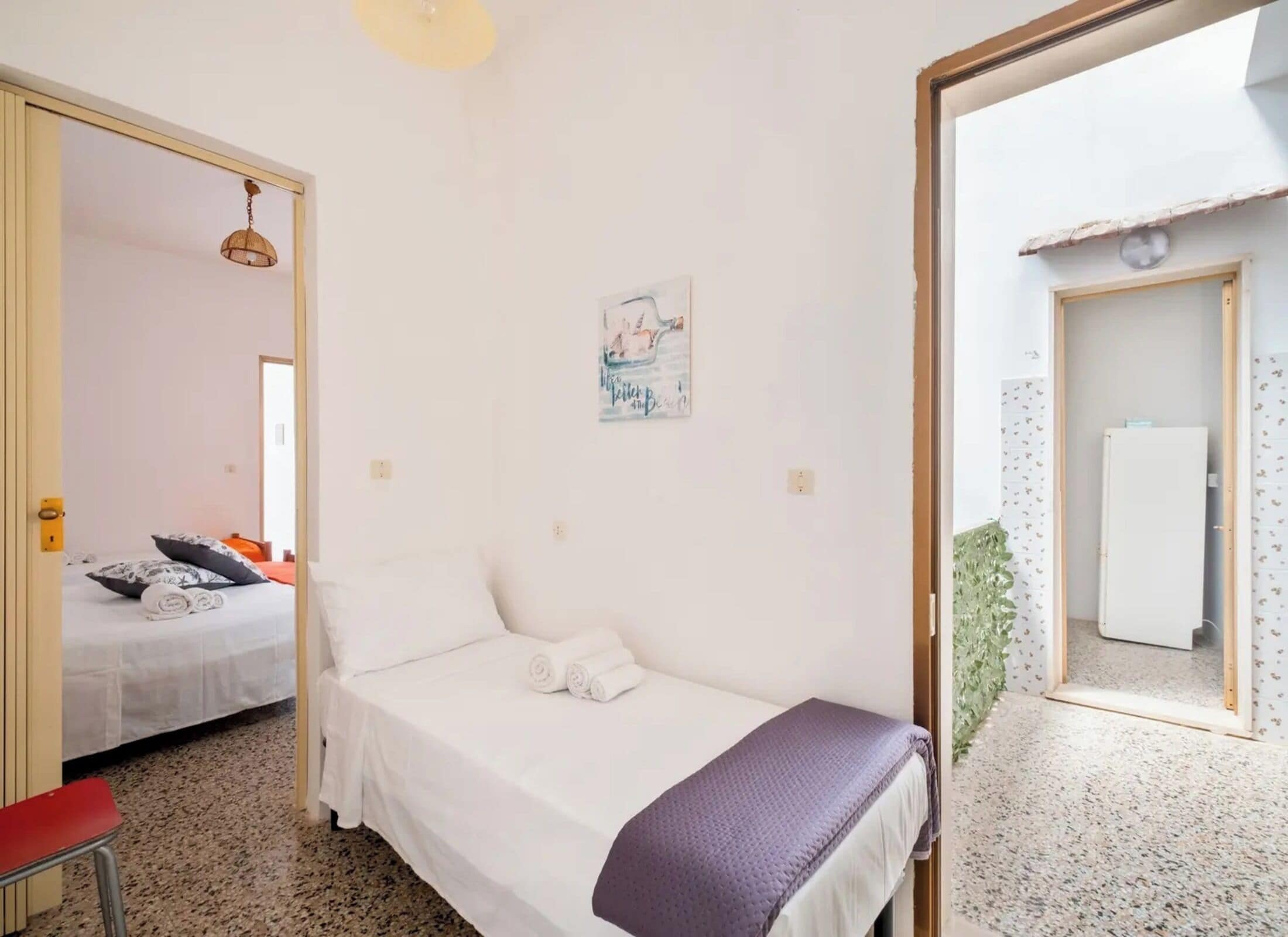 Un dormitorio con cama, una mesita, una silla, una puerta que da a otra habitación y una lámpara fijada a la pared.
