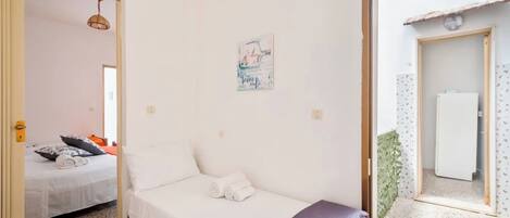 Un dormitorio con cama, una mesita, una silla, una puerta que da a otra habitación y una lámpara fijada a la pared.