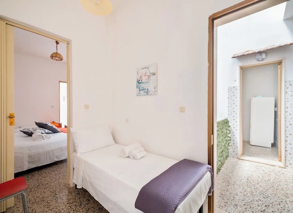 Une chambre à coucher comprenant un lit, une petite table, une chaise, une porte donnant sur une autre pièce et un luminaire fixé au mur.