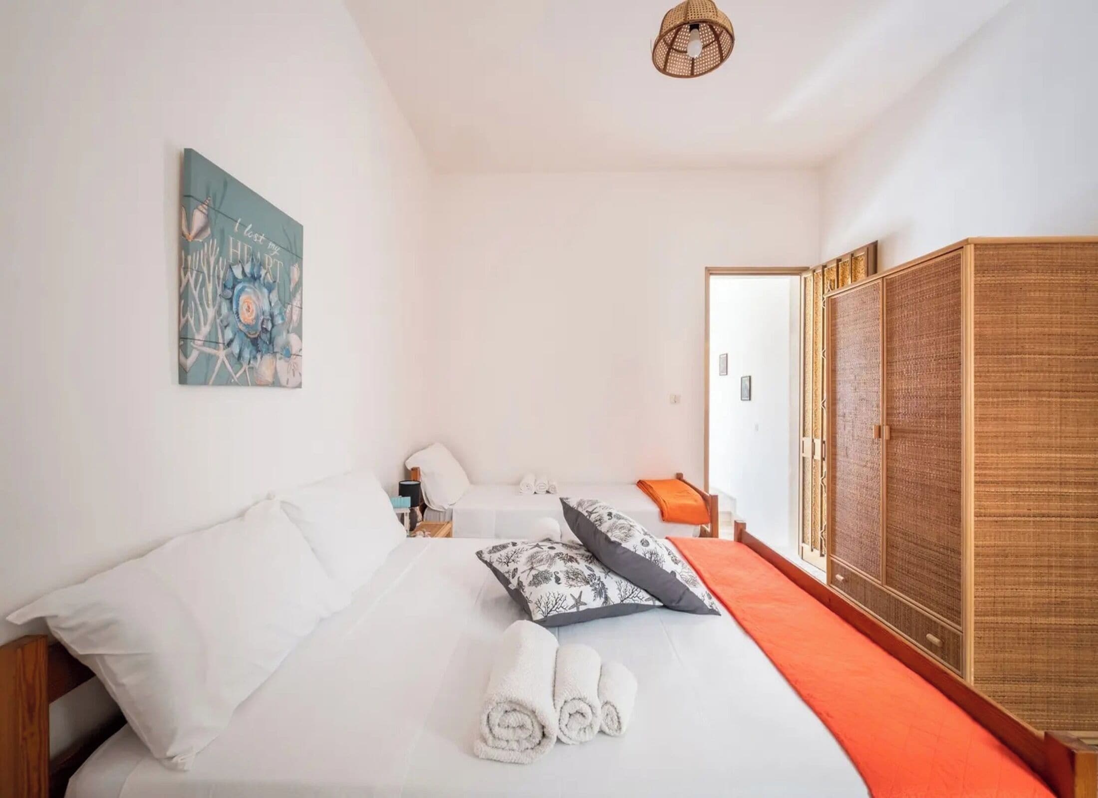 Een slaapkamer met een bed, kussens, een opgerolde handdoek, een nachtkastje, een lamp, een schilderij en een kledingkast.