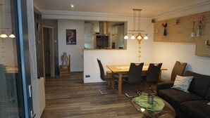 Ein modernes Wohnzimmer mit Holzboden, einem Essbereich mit Holztisch und Stühlen, einer Couch und einem Couchtisch. Im Hintergrund ist eine Küche mit Dunstabzugshaube aus Edelstahl und Pendelleuchten zu sehen.