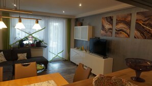 Ein modernes Wohnzimmer mit einer Couch, einem Esstisch und Stühlen. Ein Fernseher steht auf einem weißen Schrank, und an der Wand hängt ein Kunstwerk.