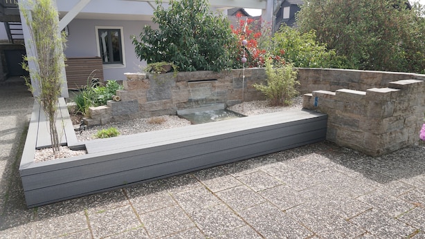 Ein moderner Garten mit einem Wasserspiel, Steinmauern und verschiedenen Pflanzen.