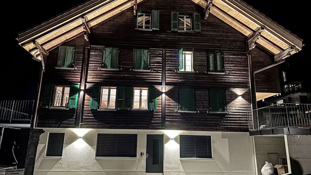A multi-story wooden building with green shutters, a balcony, and a gated entrance.