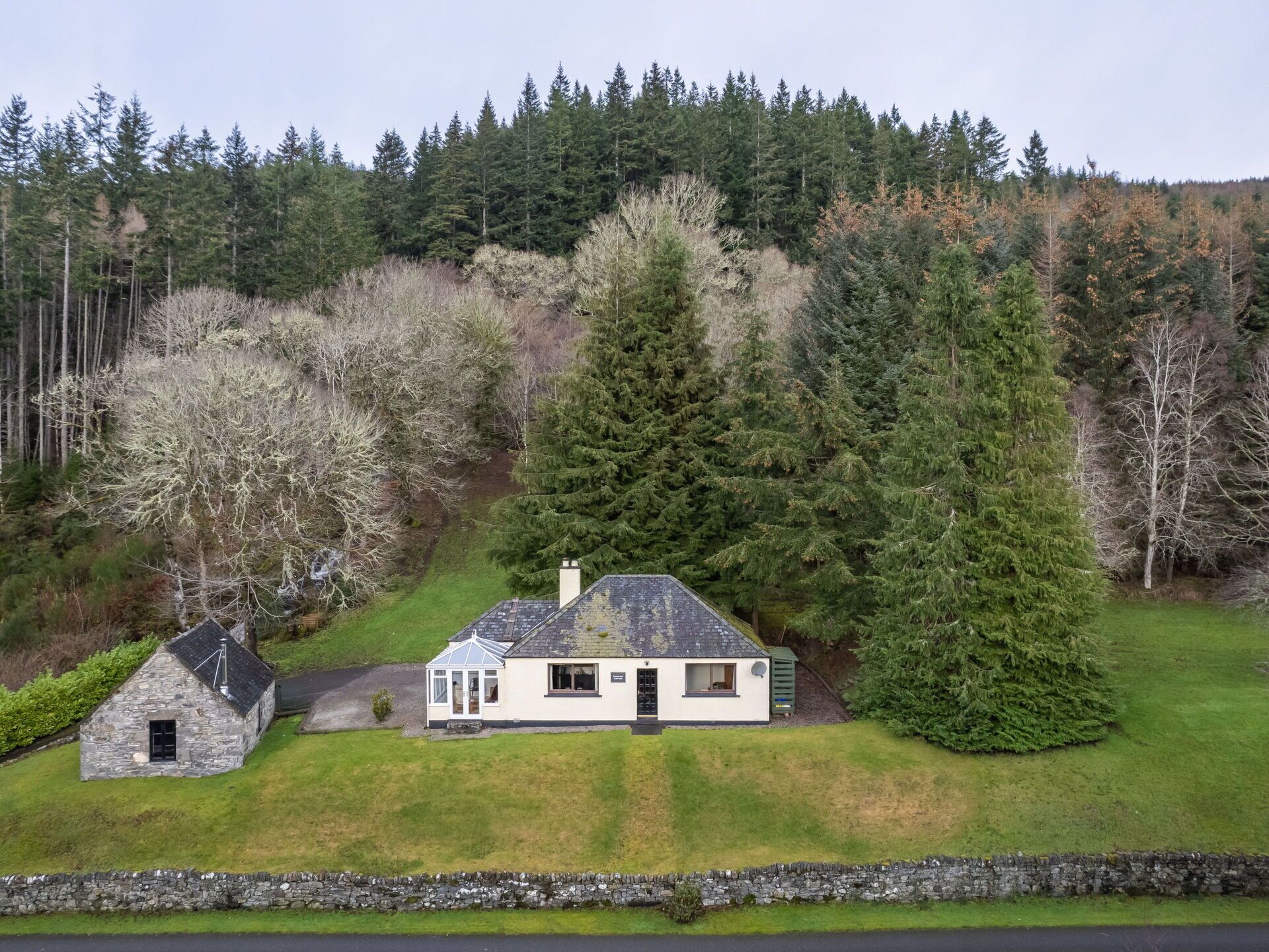 A house with a stone extension, surrounded by a lush green lawn and dense forest.
