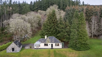 A house with a stone extension, surrounded by a lush green lawn and dense forest.