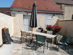 Een terras buiten met een tafel, stoelen, een barbecue en een bloempot met rode bloemen.