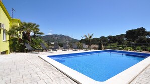 Una piscina rectangular con borde de azulejos, rodeada por un patio pavimentado con sillas de descanso.