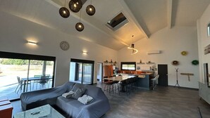 Ein geräumiger Wohnbereich mit einem grauen Sofa, einem Couchtisch und einem Essbereich mit Stühlen. Im Hintergrund befindet sich eine Küche mit einer großen Kochinsel und Pendelleuchten.