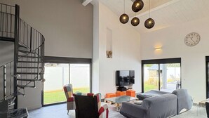 Ein geräumiges Wohnzimmer mit grauem Sofa, einem Couchtisch und einem Fernseher. Eine Treppe befindet sich links, und ein großes Fenster rechts.